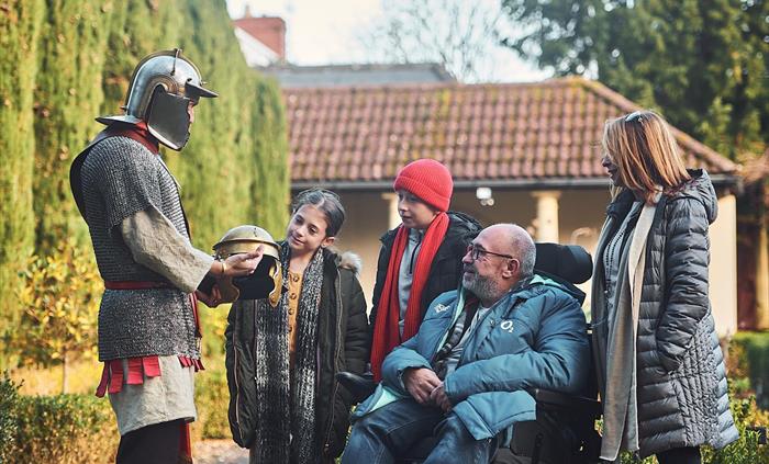 A man dressed as a Roman Gladiator talks to a family in the museum garden.